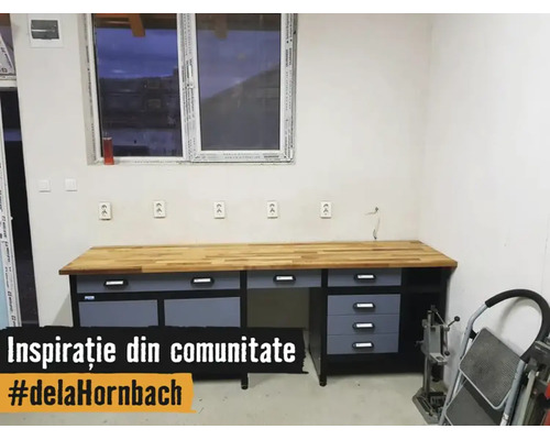 Atelier cu un banc de lucru mare, cu blat din lemn, sertare și dulapuri gri, în fața unui perete alb cu prize sub o fereastră.