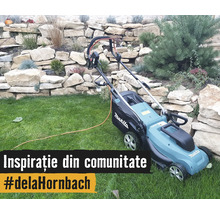 Mașină electrică de tuns gazonul Makita cu Makita Logo pe o peluză în fața unui zid de piatră cu plante în grădină.