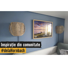Perete albastru decorat cu o ramă de tablou din lemn cu un peisaj și două aplice din împletitură naturală deasupra unui pat alb.