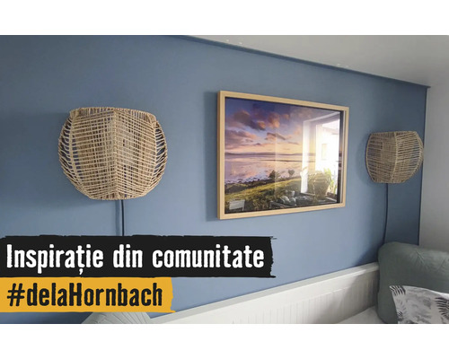 Perete albastru decorat cu o ramă de tablou din lemn cu un peisaj și două aplice din împletitură naturală deasupra unui pat alb.