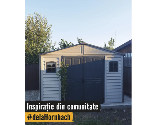 Căsuță de grădină modernă gri deschis din plastic cu uși duble gri închis și două ferestre pe o fundație de beton în grădină.