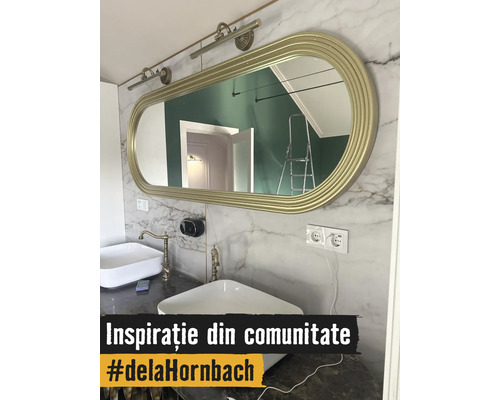 Baie modernă cu două lavoare albe, baterii aurii și o oglindă ovală mare cu ramă aurie pe un perete cu aspect de marmură.