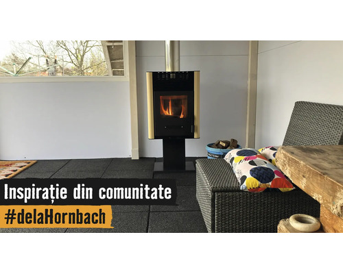 Șemineu modern pe o pardoseală neagră într-o cameră luminoasă cu mobilier de grădină din ratan și o masă din lemn rustic.