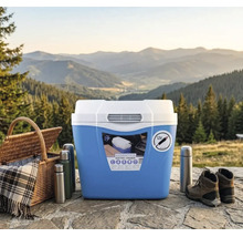 Ladă frigorifică electrică cu conexiune de 12 Volți pentru camping, lângă coș de picnic, termosuri și bocanci de munte în fața unui peisaj montan.