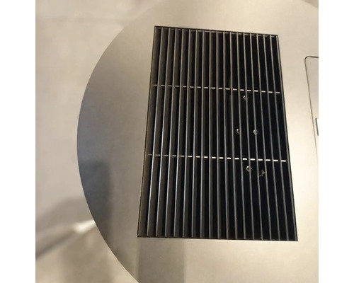 Vedere de aproape a unei grile de ventilație dreptunghiulare din metal negru, integrată într-o suprafață circulară din oțel inoxidabil.