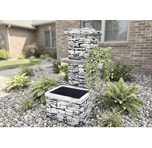 Fântână de grădină cu aspect de piatră cioplită, cu jardinieră și plante decorative într-o grădină cu pietriș.