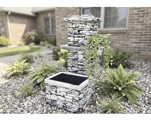 Fântână de grădină cu aspect de piatră cioplită, cu jardinieră și plante decorative într-o grădină cu pietriș.