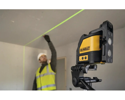 Un muncitor în construcții echipat de lucru marchează un perete folosind un laser cu linii în cruce DeWalt pe un trepied în interior.