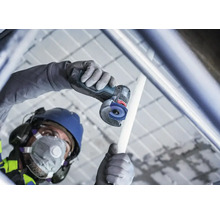 Meșter cu cască de protecție și mască de protecție respiratorie taie o țeavă cu un polizor unghiular cu acumulator pe un șantier.