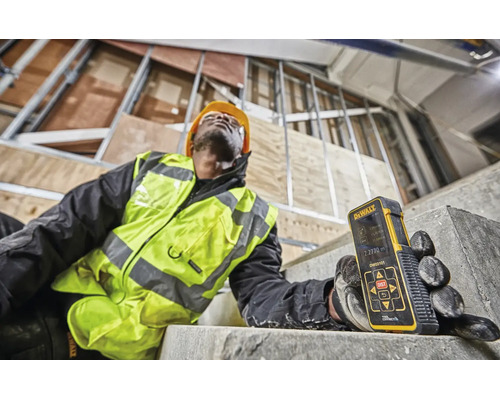 Muncitor cu telemetru laser Dewalt pe un șantier în construcție cu structură metalică și scară din beton.