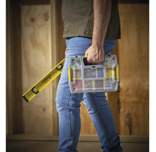 O persoană transportă o cutie de scule Stanley cu piese mici și o nivelă cu bulă de aer Stanley în fața unui perete de lemn.