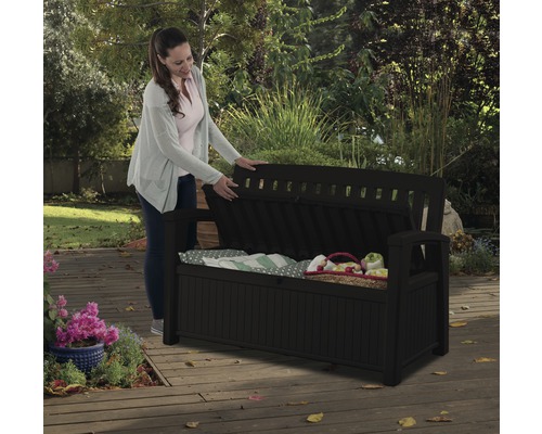 Femeie deschizând o bancă de grădină neagră cu spațiu de depozitare pe o terasă de lemn.