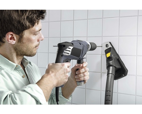 Un bărbat găurește un perete cu gresie cu o mașină de găurit lângă un aspirator