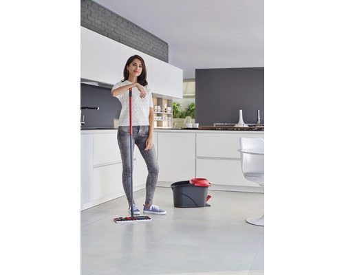 Femeie sprijinindu-se de un mop lângă o găleată într-o bucătărie luminoasă.