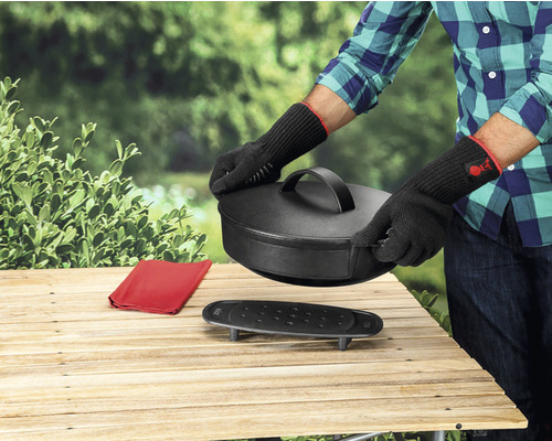 Persoană cu mănuși de grătar manevrează un cuptor olandez pe o masă de lemn în aer liber.