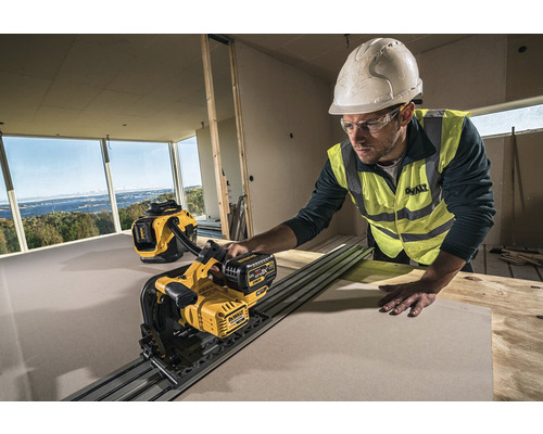 Un muncitor în construcții, cu cască și ochelari de protecție, taie o placă cu un fierăstrău pentru gips-carton DeWalt într-o încăpere în stare brută. Un aspirator DeWalt se află în fundal.