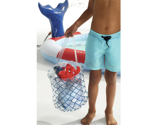 Un copil cu pantaloni de baie ține o geantă de plajă cu jucării de plajă în fața unei piscine pentru copii.