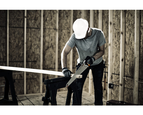 Un bărbat cu cască de protecție taie o scândură de lemn cu un fierăstrău manual pe o suprafață de lucru.