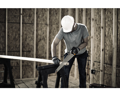 Un bărbat cu cască de protecție taie o scândură de lemn pe un banc de lucru cu un fierăstrău manual.