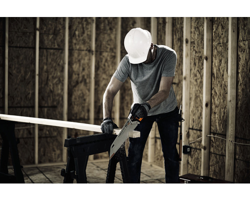 Un bărbat cu cască de protecție taie o scândură de lemn cu un fierăstrău manual pe un banc de lucru.