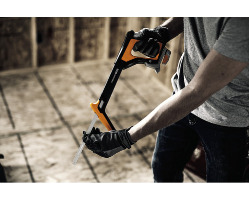 Bărbat cu mănuși și fierăstrău de mână marca Fiskars în fața unui perete de construcție brută