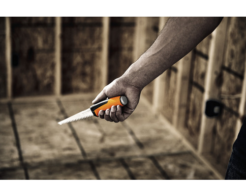 O mână ține un ferăstrău de găurit în fața unei construcții din lemn.