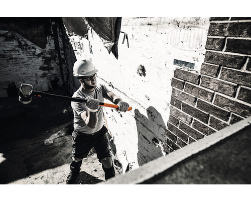 Un bărbat cu cască de protecție și ochelari de protecție lovește un zid de cărămidă cu un ciocan.