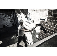 Un muncitor în construcții cu o cască de protecție lovește un zid cu un ciocan, în timp ce se află într-o încăpere care necesită renovare.