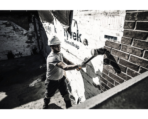 Un muncitor în construcții cu o cască de protecție lovește un zid cu un ciocan, în timp ce se află într-o încăpere care necesită renovare.