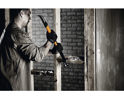 Bărbat folosind un levier pentru a demonta o structură din lemn în fața unui zid de cărămidă.