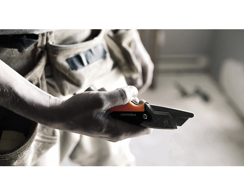 O mână ține un cuțit pentru mochetă Fiskars în fața unei curele pentru scule