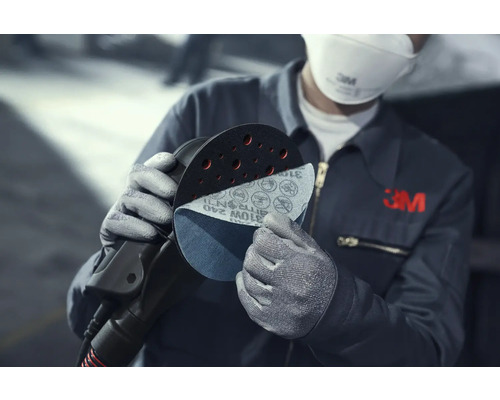 Un muncitor fixează șmirghel pe o mașină de șlefuit. Logo 3M.