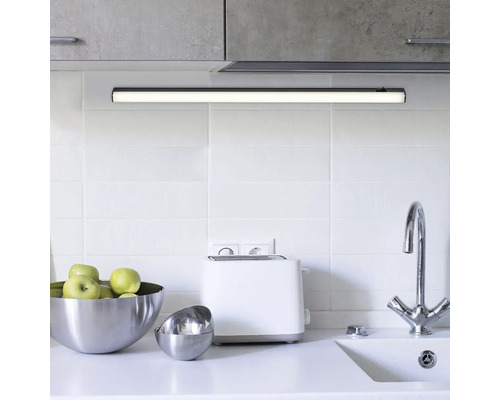 Scenă de bucătărie cu lumină LED sub dulap, chiuvetă, robinet și ustensile de bucătărie pe un blat de lucru deschis la culoare.