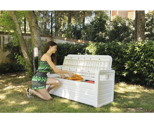 Femeie depozitând obiecte într-o bancă de grădină albă, cu spațiu de depozitare în grădină.