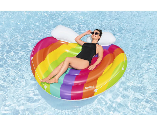 Femeie se relaxează pe o saltea gonflabilă în formă de inimă, cu motive curcubeu, în apă.