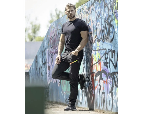 Bărbat cu tricou, pantaloni și pantofi negri, sprijinit de un zid cu graffiti