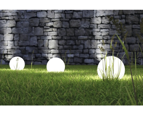 Trei lămpi sferice luminoase în grădină în fața unui zid de piatră.