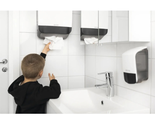 O baie cu o chiuvetă, un dispenser de prosoape și un copil care ia un prosop de hârtie.